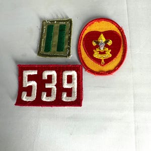May include: Three embroidered patches. One is a red rectangle with the number "539" in white. Another is a green square with two vertical green stripes. The third is a yellow circle with a red heart and a gold emblem.