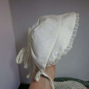 Könnte beinhalten: Ein weißes Baby-Bonnet mit Spitzenbesatz und Bändern. Das Bonnet ist aus einem weichen, strukturierten Stoff gefertigt.