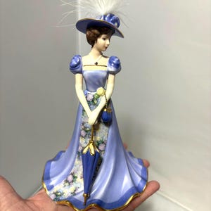 Może przedstawiać: Porcelanowa figurka kobiety w długiej, powłóczystej niebieskiej sukni i pasującym kapeluszu ozdobionym białymi piórami. Figurka trzyma niebieski parasol i małą torebkę. Suknia ma kwiatowy wzór i złote wykończenia.
