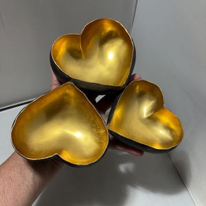 May include: Three heart-shaped bowls with a matte black exterior and a reflective metallic gold interior. The bowls are arranged in a cluster, showcasing their smooth, curved edges and deep, concave centers designed for holding small items.
