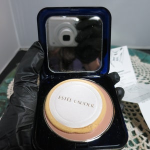 Könnte beinhalten: Ein blaues Kompakt mit einem Spiegel und einem runden, weißen, gepressten Puderkompakt mit dem Aufdruck "Estee Lauder".