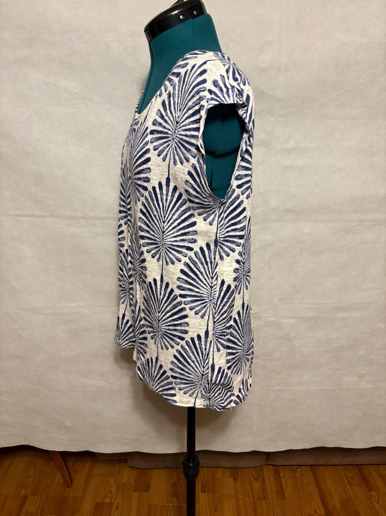 K&ouml;nnte beinhalten: Ein &auml;rmelloses Oberteil mit wei&szlig;em Hintergrund und einem sich wiederholenden marineblauen Blattmuster. Das Oberteil hat einen Rundhalsausschnitt und wird auf einer Schaufensterpuppe pr&auml;sentiert. Der Stoff scheint ein leichtes Material zu sein.