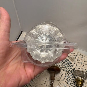 May include: A clear, faceted glass doorknob in a plastic package. The doorknob has a round shape with a decorative, ridged design. Two other vintage doorknobs are visible below. The background is a cream-colored, patterned surface.