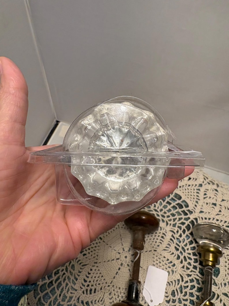 May include: A clear, faceted glass doorknob in a plastic package. The doorknob has a round shape with a decorative, ridged design. Two other vintage doorknobs are visible below. The background is a cream-colored, patterned surface.