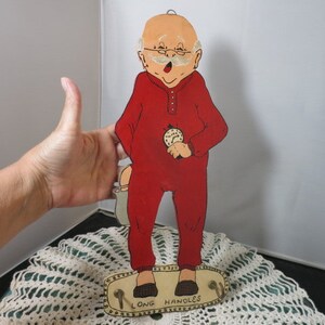 May include: A wooden cutout of a cartoon man wearing a red jumpsuit and holding a clock. The cutout is standing on a wooden base with the words "Long Handles" printed on it.