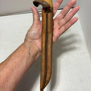 May include: A fixed-blade knife with a light-colored wooden handle encased in a tan leather sheath. The sheath features visible brown stitching along the edge and a small leather strap with a metal snap button closure at the top of the handle.