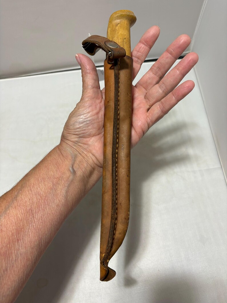 May include: A fixed-blade knife with a light-colored wooden handle encased in a tan leather sheath. The sheath features visible brown stitching along the edge and a small leather strap with a metal snap button closure at the top of the handle.
