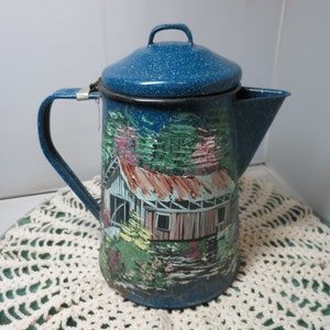 May include: Blue enamel coffee pot with a painted scene of a cabin in the woods. The pot has a lid and a handle.