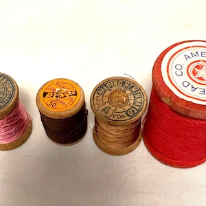 May include: Four vintage thread spools in different colours. The spools are made of wood and have labels with text. One spool is red with "AMERICAN THREAD CO." printed on the label. Other spools have pink, brown, and gold thread.