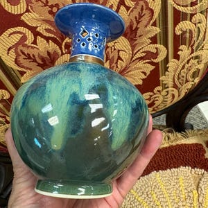 Puede incluir: Un jarrón de cerámica redondo con acabado brillante que presenta tonos mezclados de azul profundo y verde bosque. El cuello es de color azul sólido con un patrón de diamantes recortado cerca del borde, separado del cuerpo por una banda metálica.