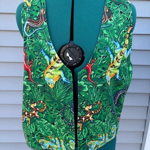 May include: A sleeveless vest featuring a vibrant jungle pattern with various lizards, including green, orange, and blue iguanas, set against a background of lush green tropical leaves and foliage. The garment has a deep V-neckline and a front opening.