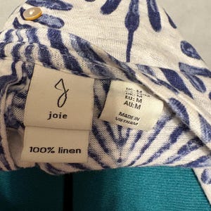 K&ouml;nnte beinhalten: Nahaufnahme eines wei&szlig;en und blau gemusterten Leinenkleidungsst&uuml;cks. Der Stoff weist ein sich wiederholendes Muster aus blauen blattartigen Formen auf. Das Kleidungsst&uuml;ck hat ein Etikett mit der Aufschrift "joie" und "100% Leinen". Zus&auml;tzliche Etiketten weisen auf die Gr&ouml;&szlig;e M und die Herstellung in Vietnam hin.