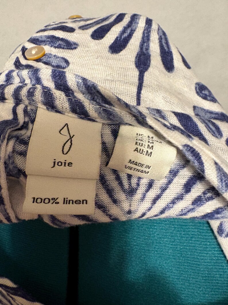 K&ouml;nnte beinhalten: Nahaufnahme eines wei&szlig;en und blau gemusterten Leinenkleidungsst&uuml;cks. Der Stoff weist ein sich wiederholendes Muster aus blauen blattartigen Formen auf. Das Kleidungsst&uuml;ck hat ein Etikett mit der Aufschrift "joie" und "100% Leinen". Zus&auml;tzliche Etiketten weisen auf die Gr&ouml;&szlig;e M und die Herstellung in Vietnam hin.
