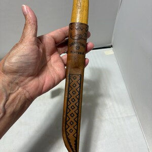 May include: A J. Marttiini knife with a light-colored wooden handle and a brown leather sheath. The sheath features a stamped fish graphic, the text J. MARTTIINI FINLAND, and a geometric diamond pattern running down the center of the leather.