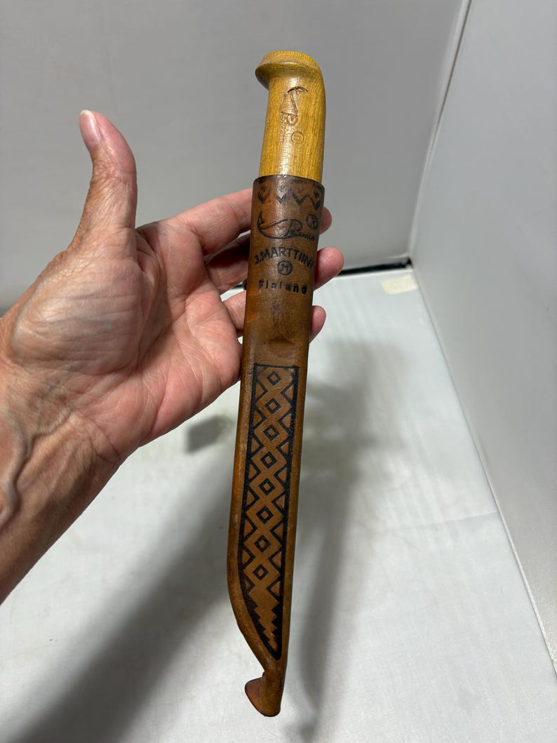 May include: A J. Marttiini knife with a light-colored wooden handle and a brown leather sheath. The sheath features a stamped fish graphic, the text J. MARTTIINI FINLAND, and a geometric diamond pattern running down the center of the leather.