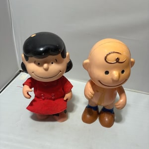 May include: Two vintage plastic figurines of cartoon characters. One figure is a girl in a red dress with black hair. The other is a boy with a bald head, brown shoes, and blue shorts. Both figures have smiling faces.
