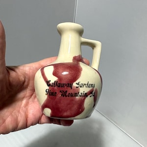 Könnte beinhalten: Ein kleiner, cremefarbener Keramikkrug mit Henkel und einem dunkelroten abstrakten Design. Der Text "Callaway Gardens Pine Mountain, Ga" ist in Schwarz auf die Vorderseite des Kruges gedruckt.