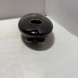 May include: A small, oval-shaped ceramic vessel with a glossy, dark brown to black glaze. The top features a single, centered circular opening with a thin, light brown rim. The base is supported by three short, rounded feet with white tips.
