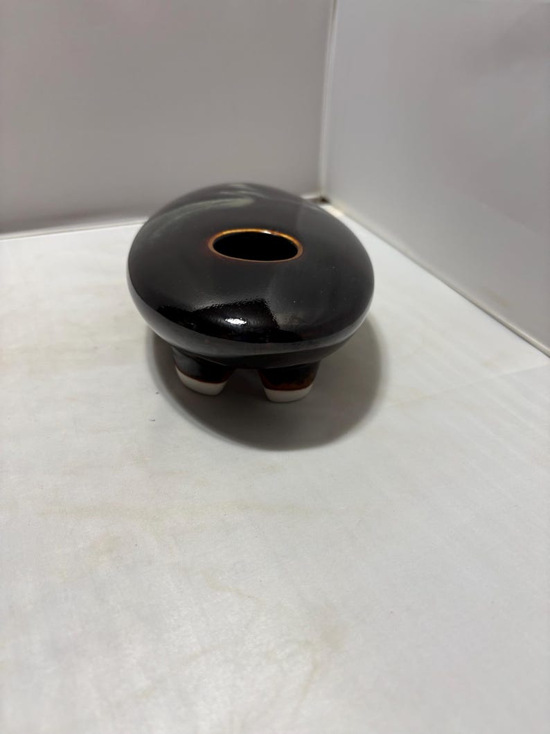 May include: A small, oval-shaped ceramic vessel with a glossy, dark brown to black glaze. The top features a single, centered circular opening with a thin, light brown rim. The base is supported by three short, rounded feet with white tips.
