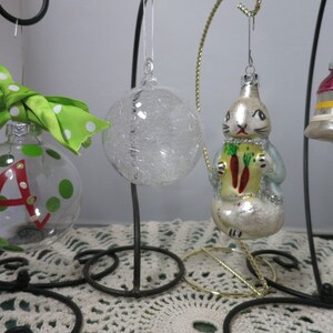 May include: Four glass Christmas ornaments hanging on a black metal stand. One ornament is clear with a textured surface, one is a white rabbit holding carrots, one is a bell with red, white, and yellow stripes, and one is clear with green polka dots and the letter "A" painted in red.