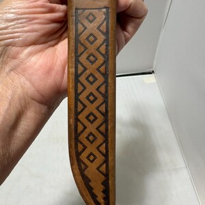May include: A tan leather sheath featuring a dark brown stamped geometric pattern consisting of a vertical row of diamonds enclosed within a border. The leather has a smooth texture and a curved shape at the bottom, showcasing detailed craftsmanship.