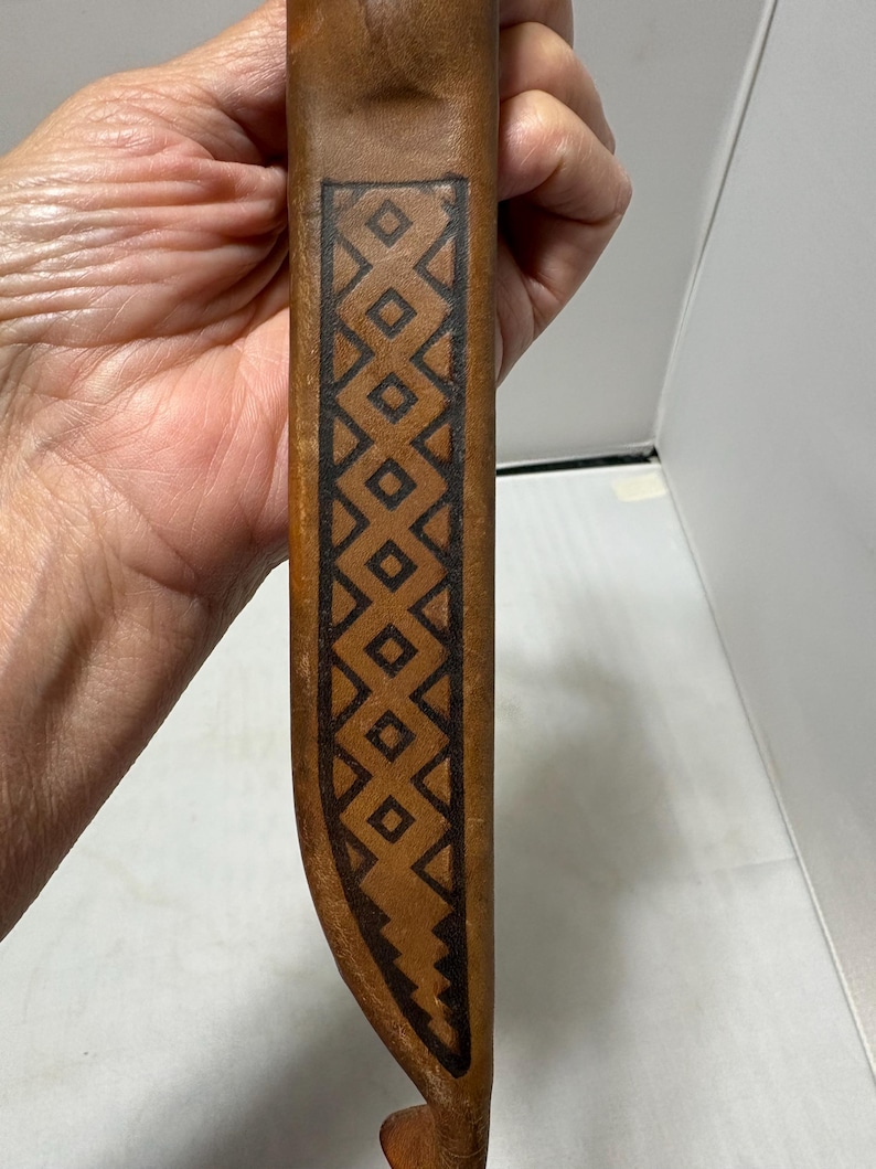 May include: A tan leather sheath featuring a dark brown stamped geometric pattern consisting of a vertical row of diamonds enclosed within a border. The leather has a smooth texture and a curved shape at the bottom, showcasing detailed craftsmanship.