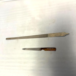 Peut inclure: Deux limes à ongles en métal. La plus grande est longue et fine, avec une extrémité pointue. La plus petite a un manche marron et le texte "TEMPLE CO. USA". Les deux limes sont de couleur argent.