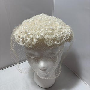May include: Ivory-colored vintage hat with a textured, curly design. The hat features a delicate net veil that drapes over the front. The hat is displayed on a white mannequin head.