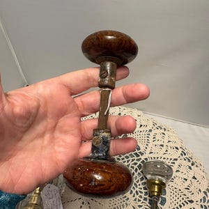 May include: Antique door knob with a dark brown, oval-shaped wooden handle. The knob is connected to a metal rod and base. The base has a dark finish with a small metal piece attached. The knob is a vintage item.