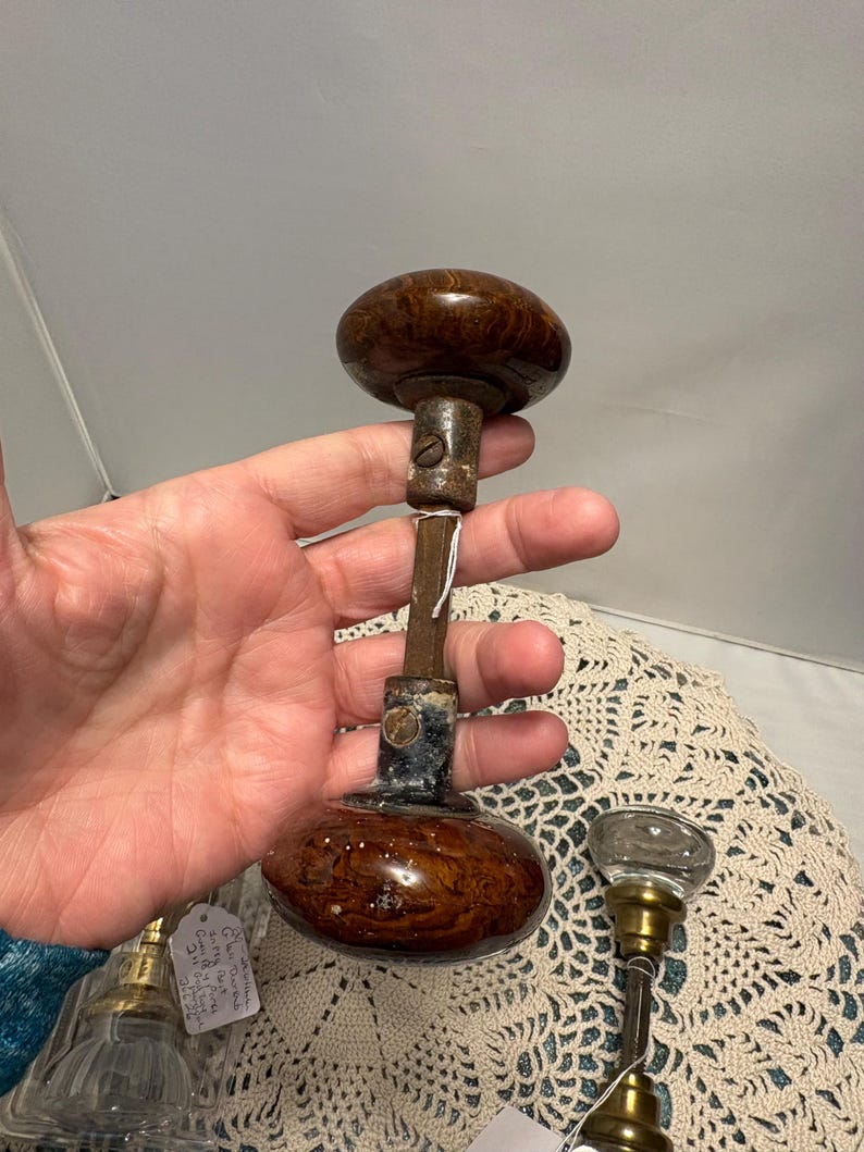May include: Antique door knob with a dark brown, oval-shaped wooden handle. The knob is connected to a metal rod and base. The base has a dark finish with a small metal piece attached. The knob is a vintage item.