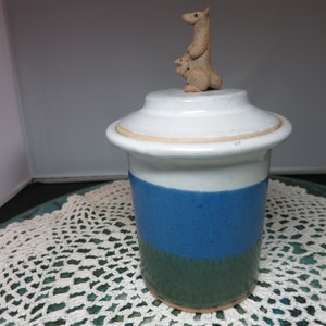 May include: A white ceramic jar with a blue and green striped design. The lid is topped with a small ceramic figurine of a kangaroo.