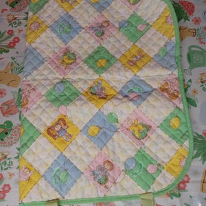 Puede incluir: Una manta de bebé acolchada con un diseño de patchwork con cuadrados pastel de color amarillo, rosa, azul y verde. Cada cuadrado tiene una imagen de dibujos animados de un bebé con un globo. La manta tiene un borde verde y correas verdes.