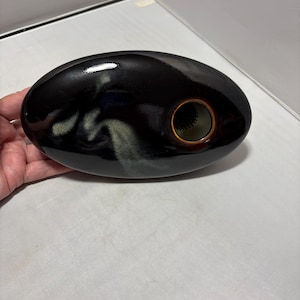 May include: An oval-shaped ceramic vase with a glossy black finish and a subtle, light-colored cloud-like glaze pattern. The vase features a single circular opening on the right side with a brown rim and a dark, spiky floral frog insert inside.