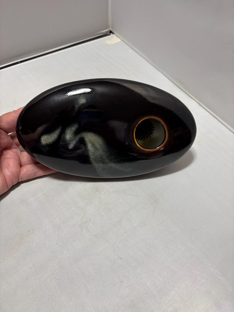 May include: An oval-shaped ceramic vase with a glossy black finish and a subtle, light-colored cloud-like glaze pattern. The vase features a single circular opening on the right side with a brown rim and a dark, spiky floral frog insert inside.