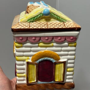 Puede incluir: Un tarro de galletas de cerámica con forma de casa de jengibre. La casa presenta un diseño de ladrillos blancos, un tejado amarillo con sombreado cruzado marrón y una puerta roja con marcos de ventanas amarillos. El tarro se sostiene en una mano.
