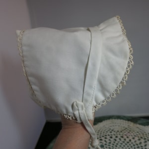 Puede incluir: Un gorro de bebé blanco con ribete de encaje. El gorro tiene un volante alrededor del borde y una corbata en la parte posterior.