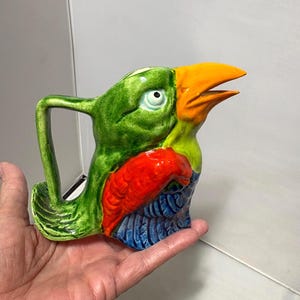 May include: A colorful ceramic bird-shaped mug with a handle. The mug features a green head, an orange beak, a red chest, and blue accents. The mug is held in a hand.