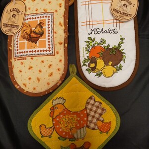 May include: Three kitchen pot holders with a chicken theme. One pot holder is yellow with a brown and white checkered pattern and a red chicken with three chicks. Another pot holder is brown with a yellow and brown floral pattern and a rooster. The third pot holder is brown with a white checkered pattern and a mushroom design. The pot holders have a brown border and a tag that says "Kitchen Compliments".