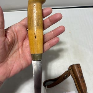 May include: A fixed-blade knife with a light-colored wooden handle and a brass bolster, featuring a straight metal blade. Beside the knife lies a brown leather sheath with a snap closure and a small white tag with handwritten text.