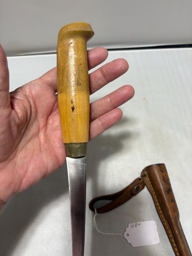 May include: A fixed-blade knife with a light-colored wooden handle and a brass bolster, featuring a straight metal blade. Beside the knife lies a brown leather sheath with a snap closure and a small white tag with handwritten text.