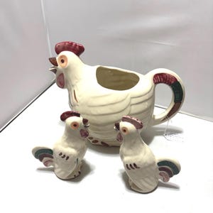 May include: A ceramic rooster-shaped pitcher and two matching rooster-shaped shakers. The pieces are cream-colored with hand-painted details in red, brown, and green. The pitcher features a large handle and an open spout, while the two smaller shakers have distinct comb and tail feather designs.