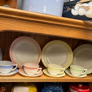 May include: A wooden cabinet shelf displays vintage pastel-colored dishware. The set includes plates and teacups with matching saucers in shades of pink, yellow, blue, and green. The plates have a ridged design.