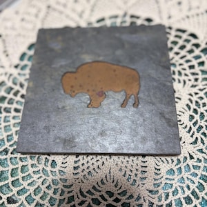 May include: Square slate coaster with a copper-colored bison silhouette. The coaster has a natural, textured surface with variations in gray and brown tones. The bison design is centered on the coaster.