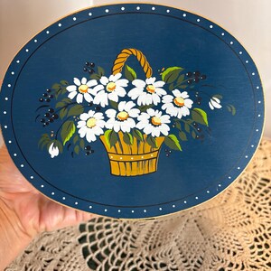 Könnte beinhalten: Eine runde Holzkiste mit einem blau bemalten Deckel, der ein handgemaltes Design eines Korbs mit weißen Gänseblümchen mit gelben Zentren und grünen Blättern auf blauem Hintergrund zeigt. Die Kiste hat einen goldenen Rand und weiße Punkte am Rand.