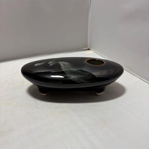 May include: An oval-shaped, low-profile ceramic vessel with a glossy black glaze and a subtle, lighter-colored cloud-like pattern on the top surface. The piece features a single circular opening and is supported by three small, short feet.