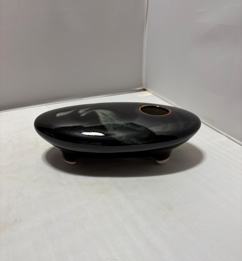May include: An oval-shaped, low-profile ceramic vessel with a glossy black glaze and a subtle, lighter-colored cloud-like pattern on the top surface. The piece features a single circular opening and is supported by three small, short feet.