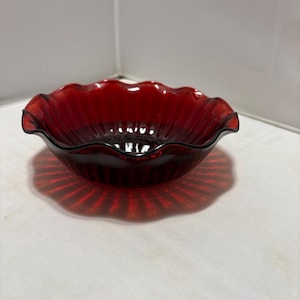 May include: A deep red glass bowl featuring a translucent finish and a scalloped, ruffled edge. The bowl has a circular shape with vertical ribbed detailing along the interior and exterior walls, creating a consistent pattern around the entire vessel.