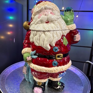 May include: A ceramic Santa Claus cookie jar. The jar is red with white trim, a black belt, and blue buttons. Santa is holding a green mitten and a blue sack. The jar is on a silver tray.