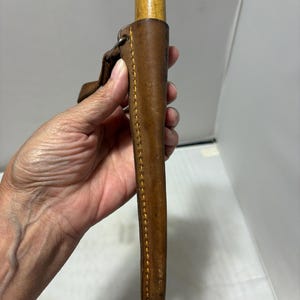 May include: A brown leather sheath with visible yellow stitching holding a tool with a light-colored wooden handle. The sheath features a metal loop attachment at the top and a tapered design that narrows toward the bottom end of the casing.