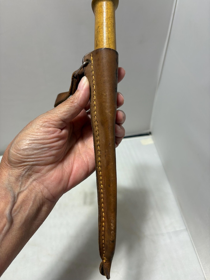 May include: A brown leather sheath with visible yellow stitching holding a tool with a light-colored wooden handle. The sheath features a metal loop attachment at the top and a tapered design that narrows toward the bottom end of the casing.